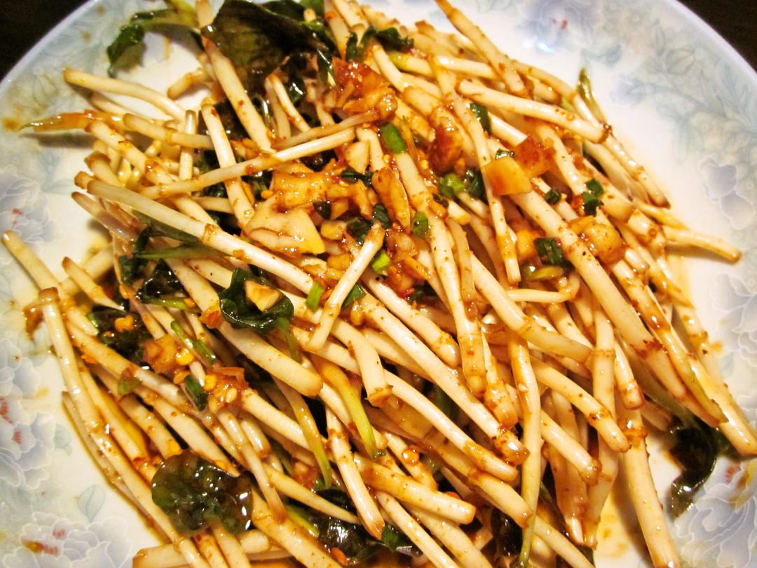 一盘晶莹翠绿的蒜薹丝凉拌菜盛放在白色瓷盘中，点缀着红色辣椒圈和芝麻，旁边放着一双木筷即将夹起一口