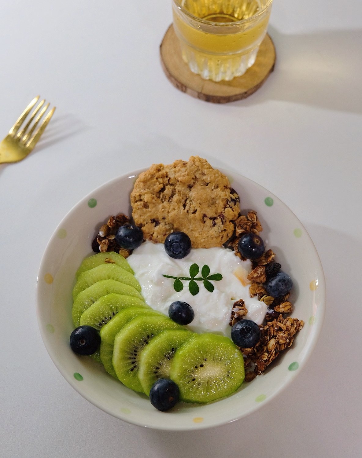 一份健康的早餐盘,包含切片的新鲜绿心猕猴桃、希腊酸奶、燕麦片、蓝莓和坚果碎,摆放在白色瓷盘中,背景为自然光线下的厨房台面,旁边有一杯绿茶和一本打开的健康食谱书 绿心猕猴桃营养早餐搭配