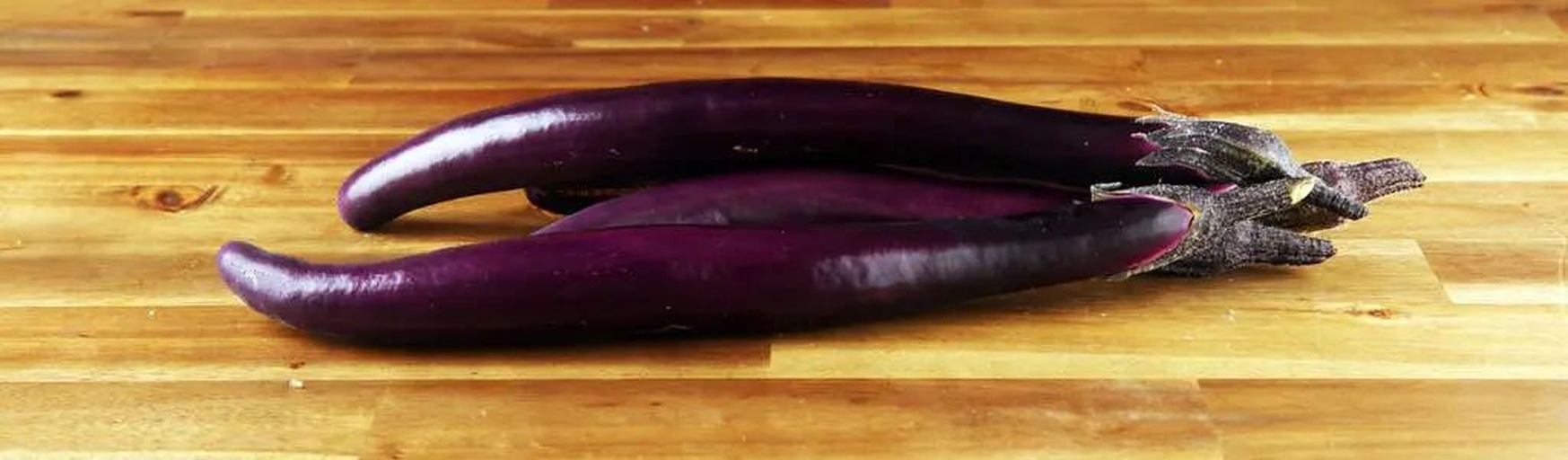 新鲜紫皮长条茄子整齐排列在左侧,右侧是装在白色小碗中的翠绿色毛豆米,背景为木质砧板和厨房刀具,光线自然明亮 新鲜茄子与毛豆米摆放在砧板上