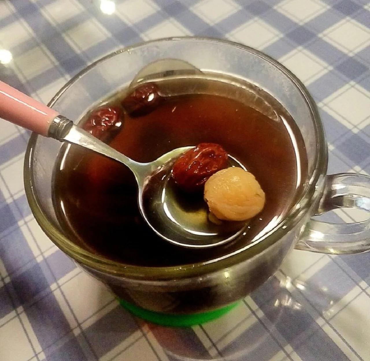 一位家庭主妇正在将干桂圆放入砂锅中,锅内已有红枣、莲子和大米,准备熬煮养生粥;台面上摆放玻璃罐装的干桂圆、量勺和竹制餐具,整体氛围温馨自然,体现日常健康饮食场景 家庭厨房中干桂圆入菜场景