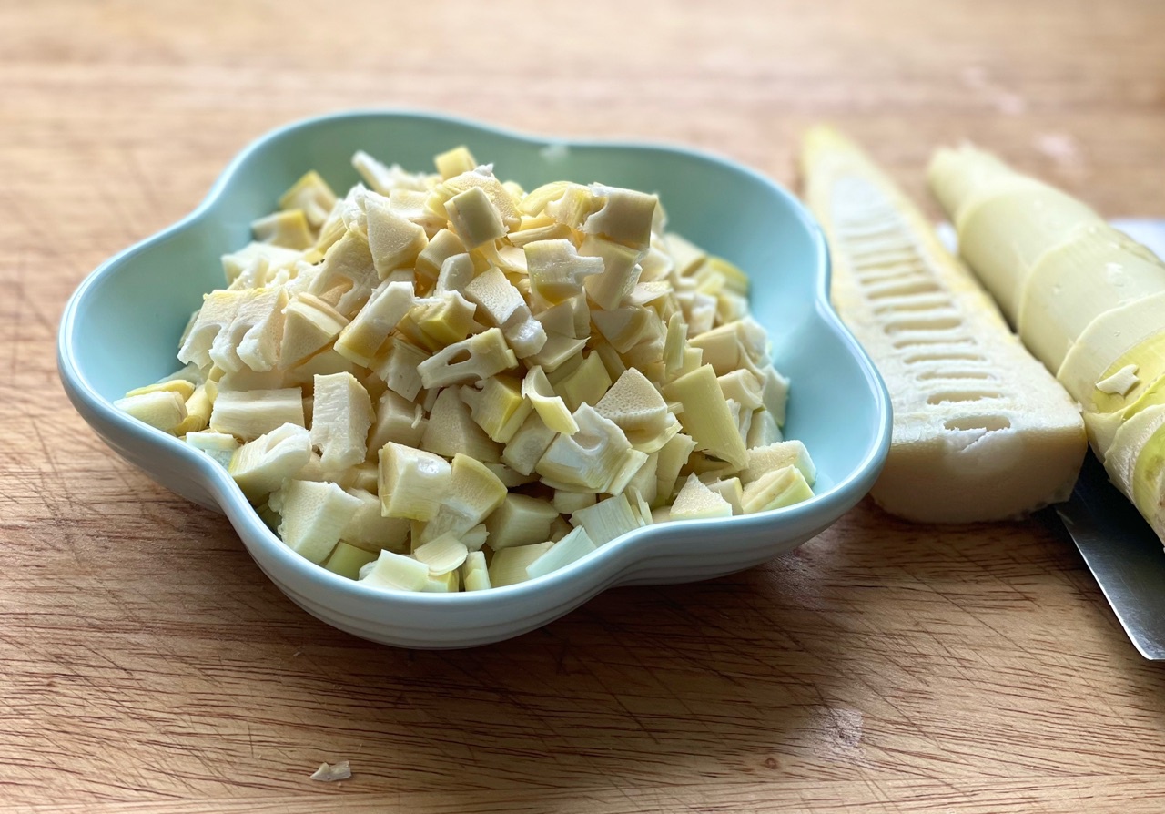 厨房操作台上摆放着一盆刚焯水冷却的竹笋块，旁边有滤网、刀具、保鲜盒和标签笔，背景可见燃气灶上的锅具