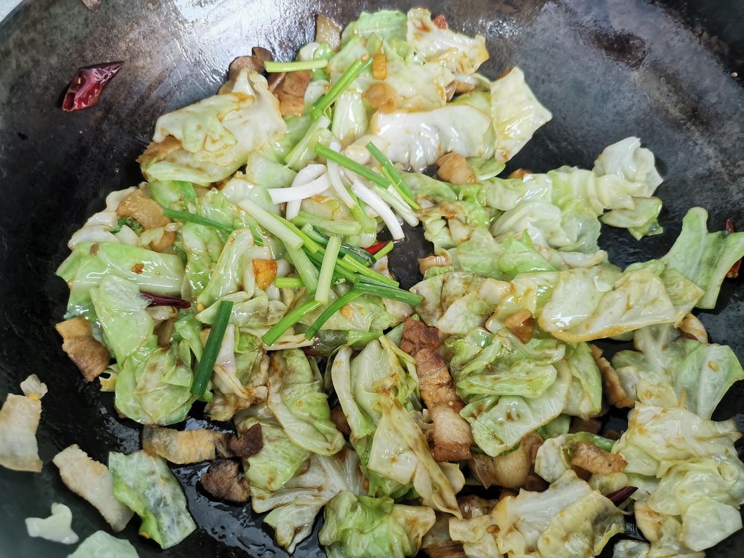 一位中年女性在明亮的家庭厨房中正在用铁锅翻炒圆包菜,锅中菜肴微冒热气,旁边放有切好的姜片和蒜瓣,台面上散落着新鲜蔬菜,展现健康家常烹饪氛围 家庭厨房烹饪场景