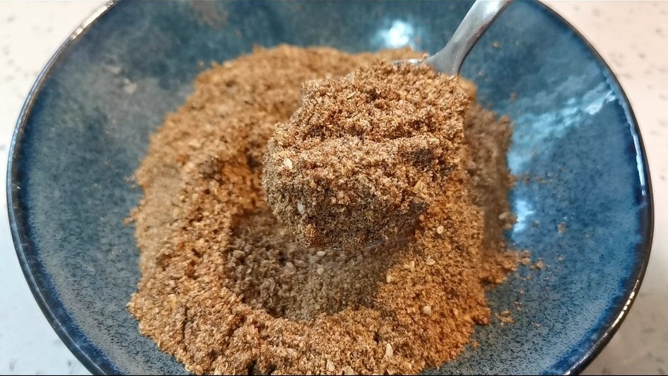 厨师正在用筷子轻柔翻拌装有切段大叶香菜和棕红色调味汁的白色瓷碗,背景可见调料瓶与芝麻罐,桌面整洁明亮 拌制过程示意图