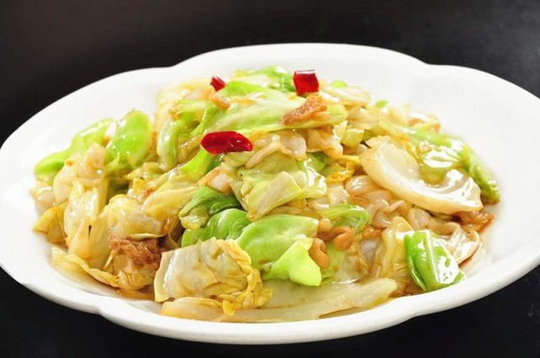 成品摆盘：一盘刚出锅的呛炒包菜，翠绿油亮，丝条根根分明，表面点缀金黄蒜末、红亮辣椒段与少许白芝麻，盛于浅口青瓷盘中，背景虚化木质餐桌