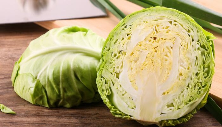 一个被横向切开的圆包菜,露出整齐的同心圆状叶层结构,颜色由外向内逐渐变浅,切面湿润有光泽,旁边摆放一把不锈钢厨刀,背景为白色瓷砖厨房台面 圆包菜切面展示