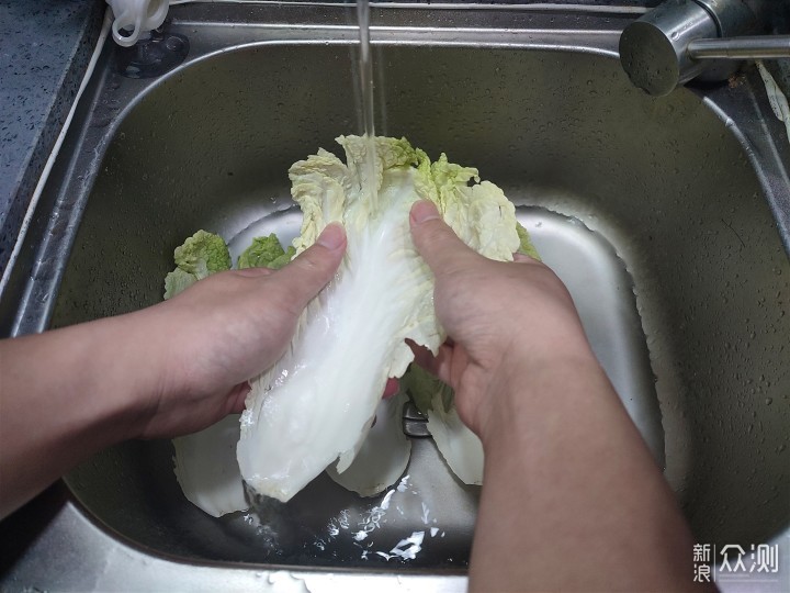 一位女性在厨房水槽中清洗大白菜,动作自然,展示实际处理黑点白菜的生活场景,桌上放有已修剪的菜帮对比样本 家庭厨房场景中,一名主妇正在清洗一颗大白菜,水流冲刷菜帮表面,旁边摆放着切掉黑点后的菜帮样本