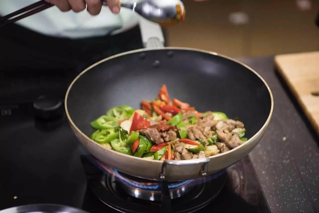 一位身穿白色厨师服的厨师正在现代化厨房中翻炒锅中的食材,锅中可见清晰的切段线椒与肉类混合,热气腾腾,旁边案板上摆放着新鲜的整根线椒,背景为不锈钢厨具与悬挂的炊具 厨房中厨师使用线椒烹饪场景