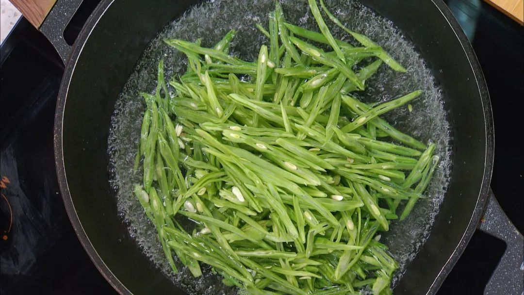 一位厨师正在将切好的新鲜荷兰豆倒入沸腾的加盐水中进行焯烫,旁边放置一碗冰水用于快速冷却,台面上有干净的砧板、刀具和已处理好的豆荚,背景为现代厨房环境 荷兰豆焯水与冷却过程示意图