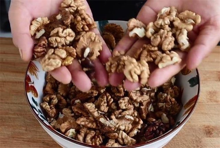 家庭场景核桃科学食用组合：左上角为刚剥出的完整核桃仁（浅琥珀色、表面油润有光泽），右下角配新鲜猕猴桃切片与红椒条，中间用陶瓷小碟分隔，背景为木质餐桌与食品级不锈钢夹