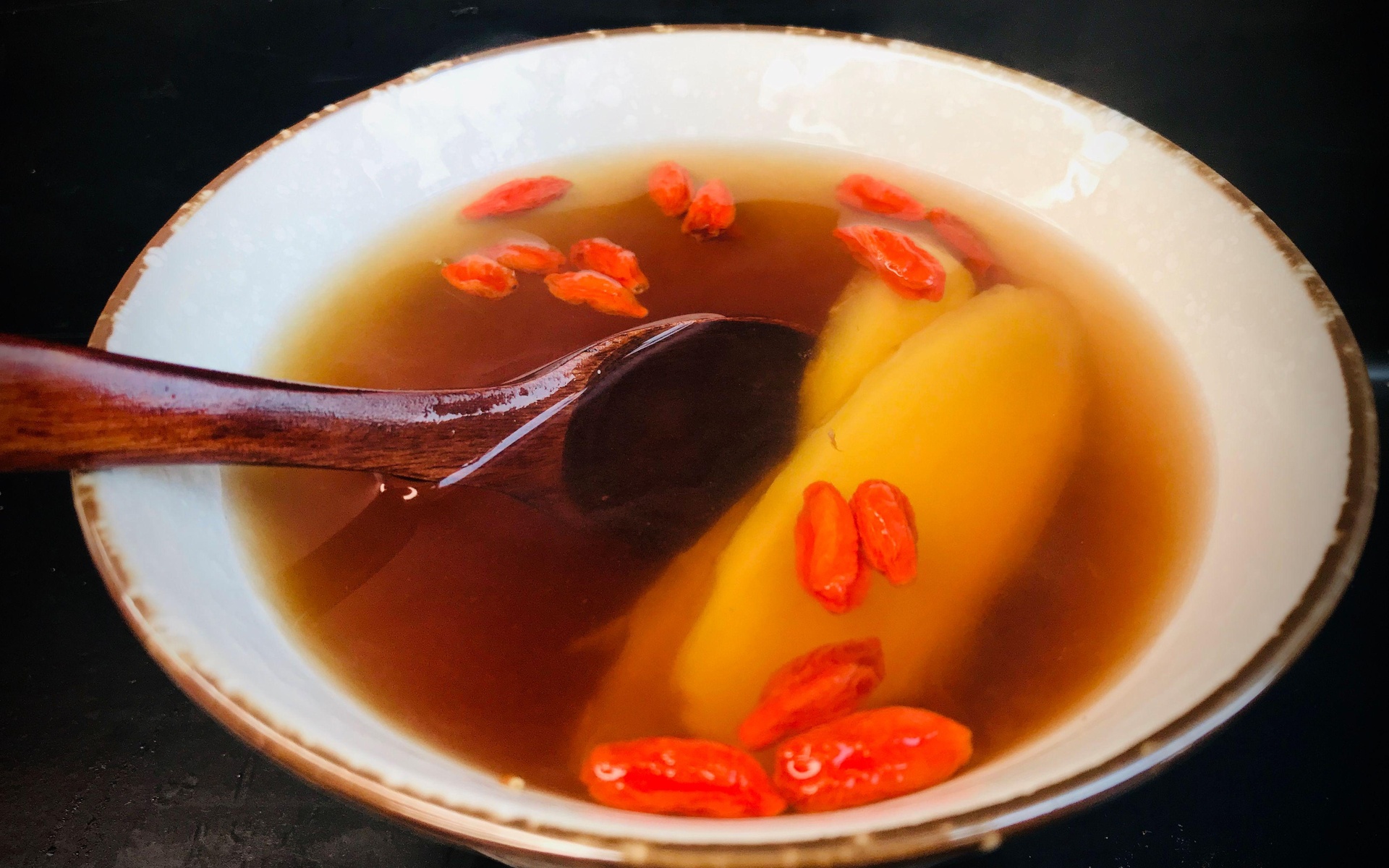 一碗刚出锅的蒸香蕉,香蕉呈深黄色,质地柔软,表面微微反光,碗边点缀着几颗红色枸杞,旁边放有一把陶瓷勺子,背景为浅色木质餐桌 成品蒸香蕉特写