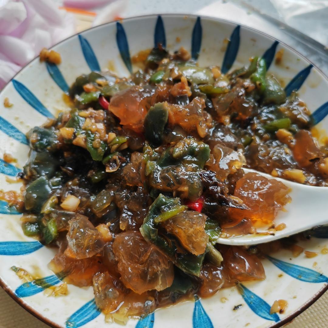一道精致的烧椒皮蛋摆放在白色瓷盘中,皮蛋切成六瓣均匀分布,周围环绕着手工撕成条状的深绿色烤椒,表面泛着微微油光。点缀有细碎蒜末和红油亮泽的调味汁,旁边配有几片香菜叶装饰。整体色彩对比鲜明,展现川菜特有的视觉冲击力与食欲诱惑 烧椒皮蛋菜品特写图