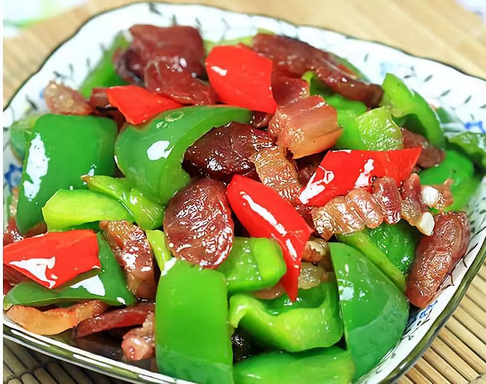 青、红、黄三色圆椒片与琥珀色广式腊肠段均匀分布于白瓷圆盘中,椒片饱满挺括、色泽鲜亮,腊肠油润微卷,表面泛自然光泽,无汁水析出,背景为木质餐桌与竹制饭勺 腊肠炒圆椒成品特写