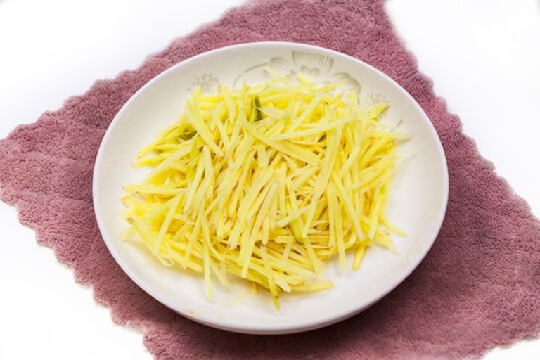 一位厨师正在厨房操作台上用刀将黄色嫩姜切成细丝,旁边有一碗清澈的冰水,姜丝已部分浸入水中,背景为木质砧板和不锈钢调料罐,光线自然明亮 面姜切丝与冰水浸泡过程