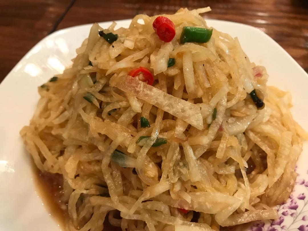 一位家庭主妇正在明亮的厨房中用刀将青萝卜切成细丝,案板旁放有酱油瓶、醋瓶、蒜瓣和香油壶,操作台上有已完成的凉拌青萝卜成品,颜色翠绿洁白相间,点缀着红辣椒丝,整体呈现健康家常菜风格 家庭厨房中制作凉拌青萝卜的场景