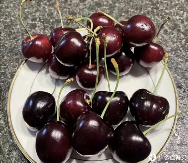 一盘新鲜的深红色车厘子,果实饱满圆润,表面有光泽,背景为浅色木桌,旁边配有绿色叶片和透明玻璃碗,展示车厘子作为高档水果的视觉形象 车厘子特写照片