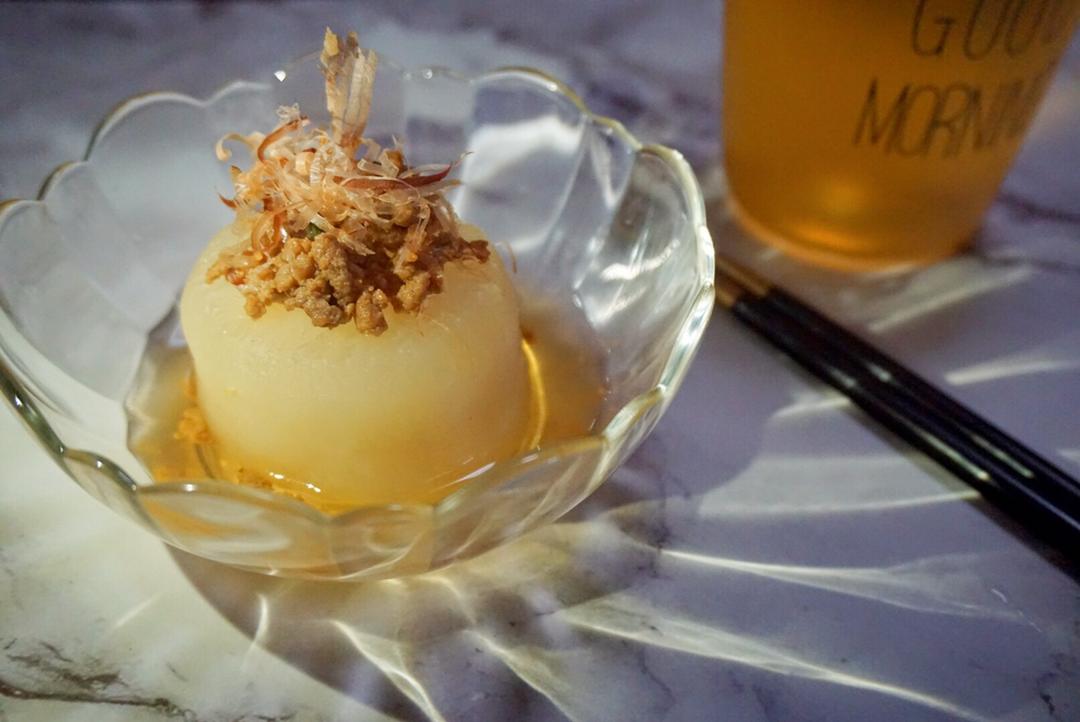 关东煮炖煮过程图,展示白萝卜在清汤中炖至半透明的状态,汤面漂浮少量木鱼花,背景为木质餐桌 一锅正在慢炖的关东煮,白萝卜块半透明状,漂浮在琥珀色清汤中,锅边配有昆布和木鱼花原料