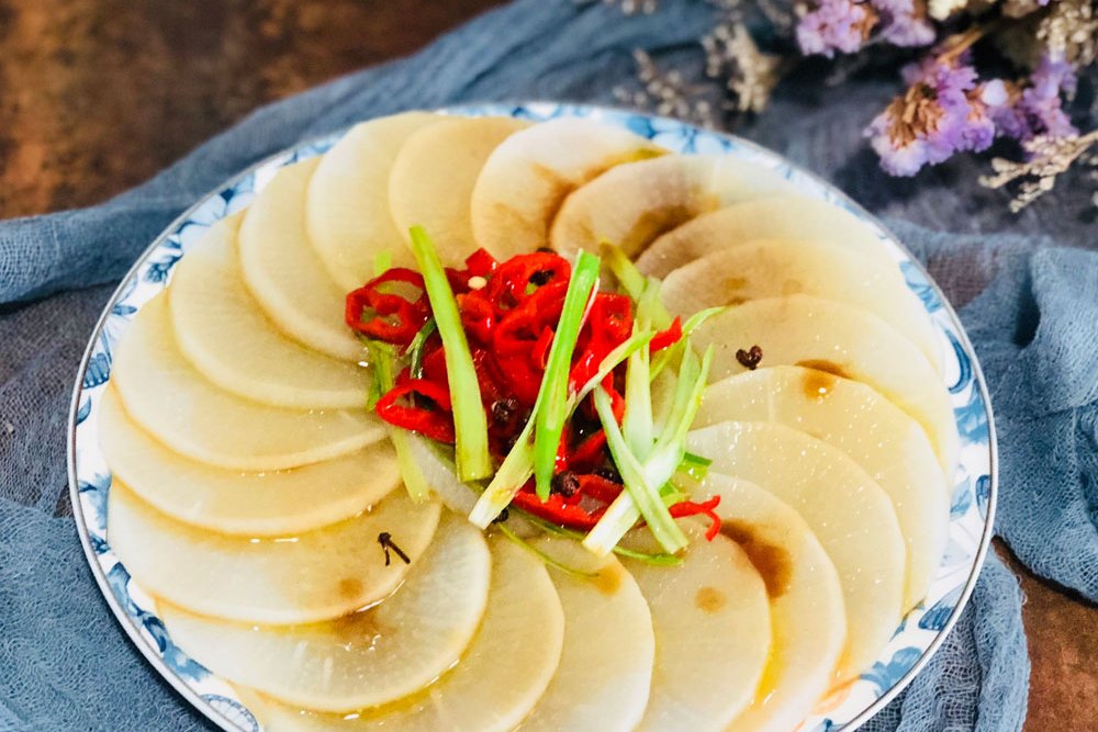 温馨的家庭厨房场景,一位主妇正在案板上用刀将青萝卜切成薄片,旁边放着酱油瓶、辣椒油和香菜碗,准备制作凉拌青萝卜。桌上还有一盘已完成的凉拌菜,青萝卜片晶莹剔透,点缀着红椒丝和香菜,整体色彩鲜明,充满生活气息,展现青萝卜的常见食用方式 家庭厨房中使用青萝卜制作凉拌菜的场景
