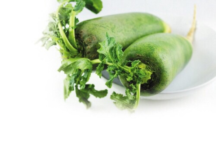 一根新鲜的青萝卜,表皮光滑呈翠绿色,顶部带有绿色叶子,根部略微泛白,放置在木制砧板上,背景为自然光下的厨房环境 青萝卜特写