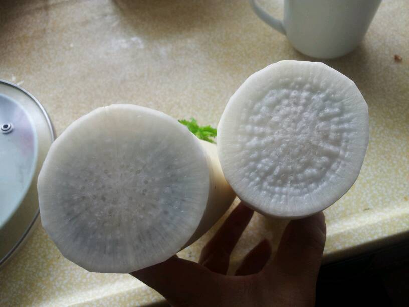 一位家庭主妇在明亮的厨房中用刀将白萝卜和胡萝卜分别切成薄片,案板旁放着两种蔬菜的整根样本,背景有锅具和调料瓶,体现日常烹饪生活氛围 家庭厨房中切萝卜的场景