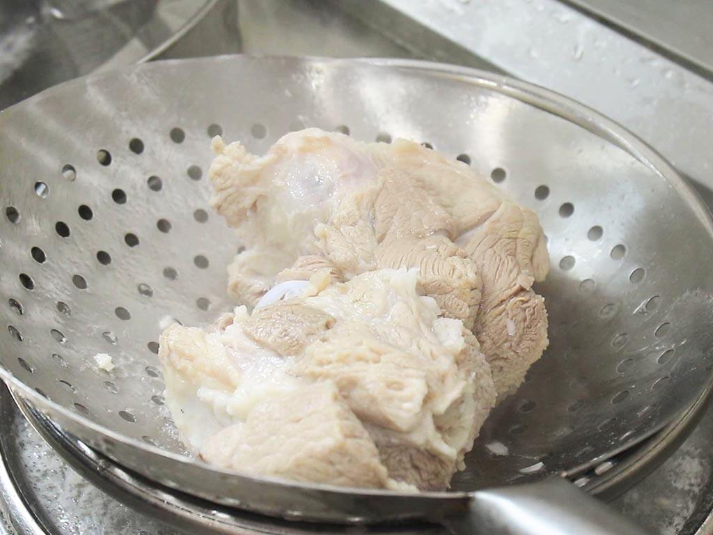 不锈钢锅中热水翻滚,瘦肉块正在焯水,表面浮起白色泡沫,一只手拿着漏勺准备捞出肉块,旁边摆放着切好的姜片和料酒瓶,厨房背景整洁明亮 红参瘦肉焯水过程