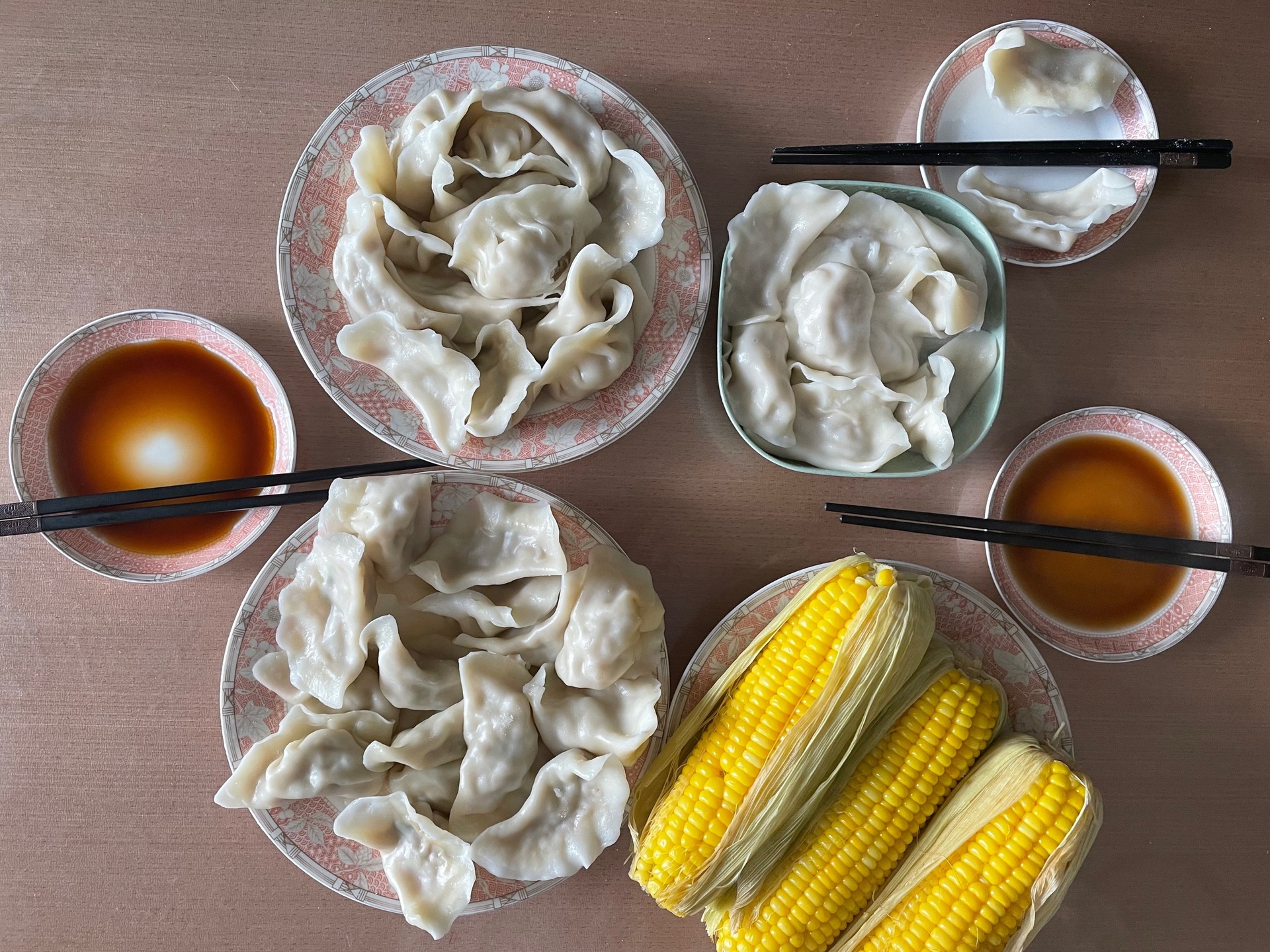一盘刚出锅的玉米馅饺子整齐摆放在白色瓷盘中,饺子呈半透明状,能隐约看到内部金黄色的玉米粒,表面略带油光,旁边配有小碟蘸料,包含酱油、醋和红油,背景为木质餐桌 蒸好的玉米馅饺子摆盘图