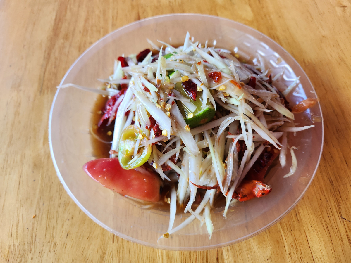 一碗色彩丰富的泰式青木瓜沙拉,内含刨成细丝的青绿色未成熟木瓜、小番茄、豇豆段、花生碎和辣椒,淋有酱汁,背景为热带风格餐布,体现异域风味料理场景 泰国青木瓜沙拉特写图