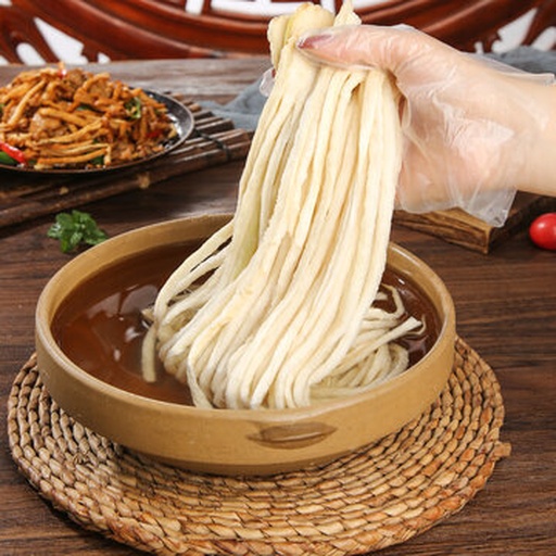 家庭厨房实操场景：戴围裙的厨师正用长筷将鱼片轻柔滑入沸腾红汤，灶台左侧摆着切好的西红柿块与酸菜丝，背景可见不锈钢锅与木质料理台