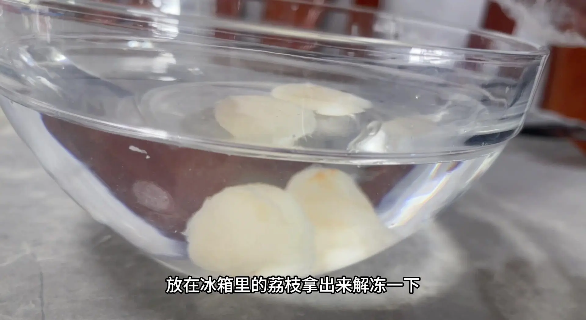 一个黑色冻梨浸泡在透明玻璃碗的清水中,水面微微泛起气泡,冻梨表面有细微冰晶融化痕迹,背景虚化的厨房环境显示为冬季室内,窗外可见薄雪,强调‘冷水缓化’的真实场景 冻梨解冻过程特写