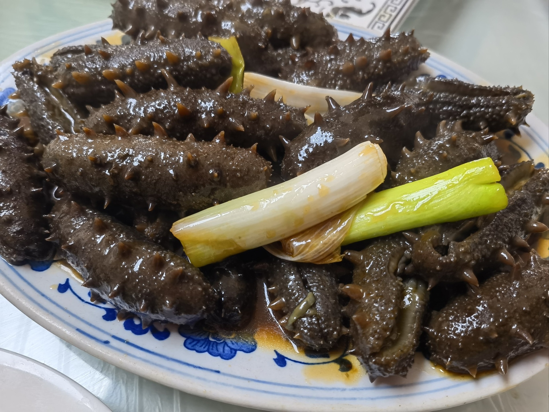 一位厨师正在不锈钢灶台上用铁锅烹制葱烧海参,锅中可见整段鸡腿葱与海参同烧,汤汁浓稠红亮,鸡腿葱段完整未切断,突出其长葱白特性。背景有调料瓶、砧板上剩余的鸡腿葱原料 厨房中正在用鸡腿葱烹饪葱烧海参的场景