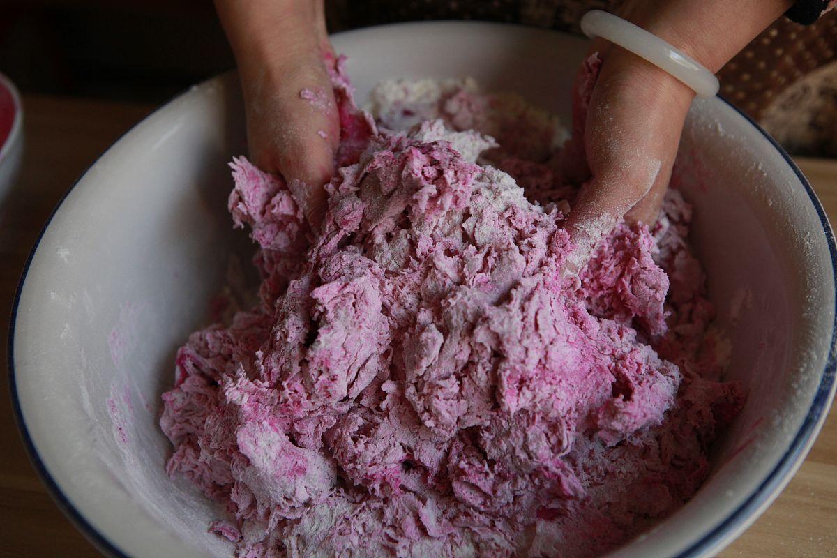 两名身穿白色厨师服的师傅正在工作台上对一个巨大的白色面团进行手工塑形,面团已初具桃子轮廓,一人用刮刀修整边缘,另一人用小刷子涂抹红色色素于顶部 巨型寿桃手工塑形现场