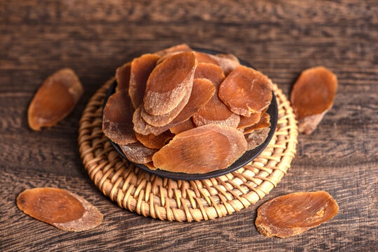 一位中年女性正在厨房将鲜人参切片放入炖盅,旁边摆放着枸杞、红枣;另一侧桌上放有红参片和研磨器,背景为中国传统风格的橱柜和药材罐子 家庭食用人参场景图