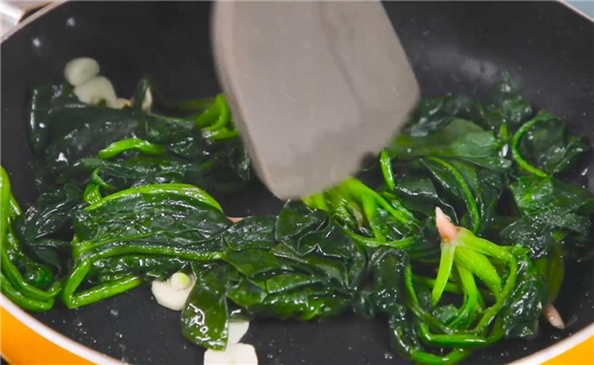 厨房操作台上的炒锅内正在翻炒菠菜与咸鸭蛋黄,可见蛋黄呈沙状包裹菠菜叶,旁边摆放剥壳的咸鸭蛋和洗净的菠菜原料 图片描述
