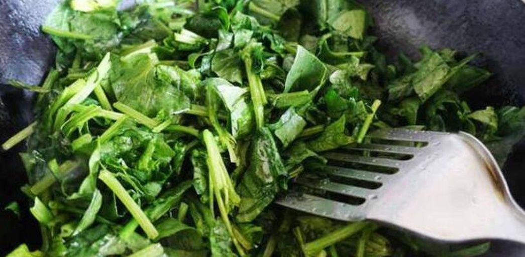 一位厨师在明亮的家庭厨房中用铁锅翻炒大叶菠菜,锅中蔬菜呈翠绿色,配有蒜瓣和少量红椒丝,灶台旁摆放着未处理的整株菠菜和砧板,体现健康家常菜制作过程 家庭厨房中烹饪大叶菠菜的场景