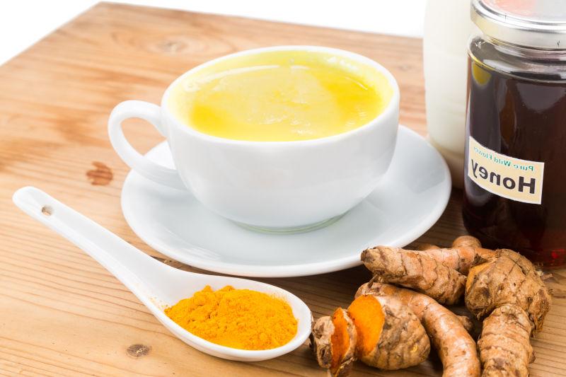 一张白色木桌上摆放着三杯不同形态的黄姜饮品:一杯是金黄色的温热黄金奶,杯口飘着蒸汽;一杯是透明玻璃瓶装的黄姜柠檬蜂蜜水;第三杯是黄姜粉末与温水混合的冲剂,旁边散落几粒黑胡椒和一小勺蜂蜜,整体呈现自然健康的生活方式美学 黄姜制成的养生饮品展示