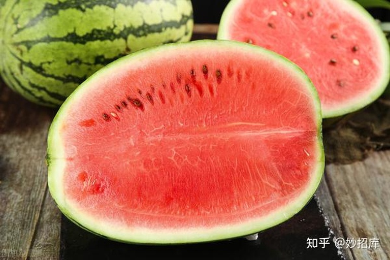一颗完整的红瓤拇指西瓜放置在白色背景上,瓜体呈椭圆形,表皮为翠绿色带有深绿纵向条纹,一旁切开的半颗显示内部鲜红色果肉,汁水饱满,果肉细腻无明显纤维,旁边配有标准尺子显示长度约为7厘米 红瓤拇指西瓜特写