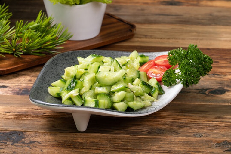 一名年轻女性穿着围裙在明亮现代厨房的操作台上用刀将一根翠绿色的小黄瓜切成薄片,旁边摆放着橄榄油瓶、柠檬和生菜叶,准备制作健康蔬菜沙拉 一位女性在厨房中将新鲜小黄瓜切片放入玻璃碗中制作沙拉