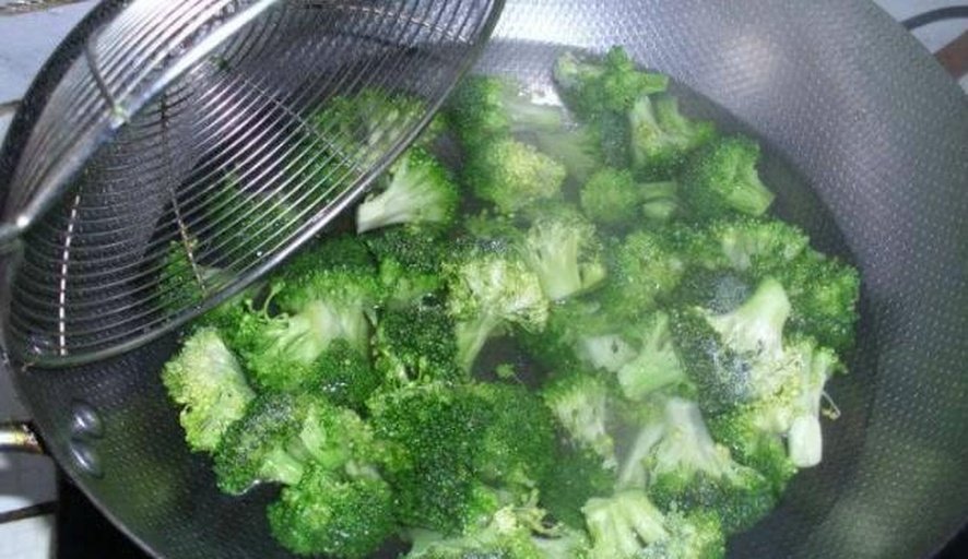 西兰花被分成小朵，正在沸水中焯烫，锅边有蒸汽升起，旁边放着一碗冷水用于快速降温