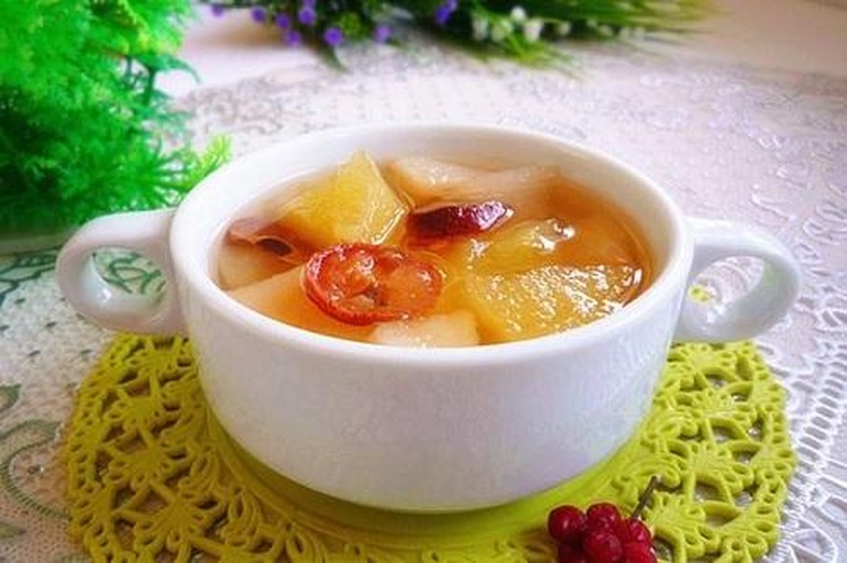 一杯清澈金黄色的香梨水被缓缓倒入透明玻璃杯中,杯壁透出温暖光泽,旁边放置一朵干百合和几粒枸杞,背景为木质餐桌与自然光环境,整体氛围温馨养生 成品香梨水倒入玻璃杯中的画面