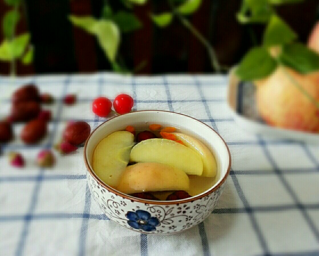 一杯热腾腾的苹果大枣枸杞水放置在木质餐桌上,旁边有食材原料点缀 苹果大枣枸杞煮水成品图