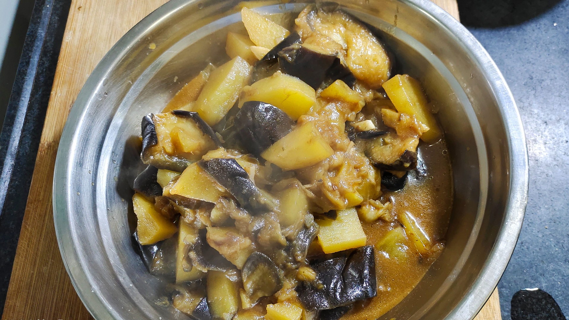 一碗刚出锅的排骨土豆茄子炖菜特写,红褐色汤汁浓郁光亮,排骨色泽油润,土豆块分明,茄子条软糯,表面撒有绿色葱花,旁边配有一碗白米饭,整体构图突出食物的食欲感与家庭温馨氛围 图片描述