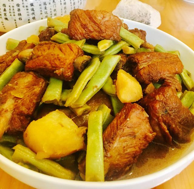 一口铸铁锅中正咕嘟咕嘟炖着排骨与白不老豆角,汤汁浓稠微沸,豆角已呈现出半透明软糯状态,排骨色泽红亮,蒸汽氤氲,体现家常炖菜的温暖氛围 锅中正在炖煮的排骨炖白不老豆角