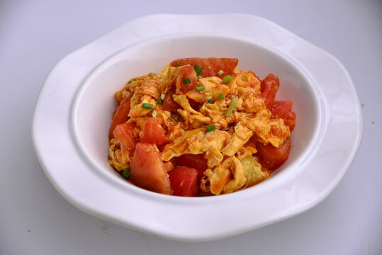 成品柿子炒鸡蛋盛放在白色瓷盘中，蛋块金黄、柿子鲜亮，点缀少许葱花，背景为简约餐桌布置，营造家庭用餐氛围