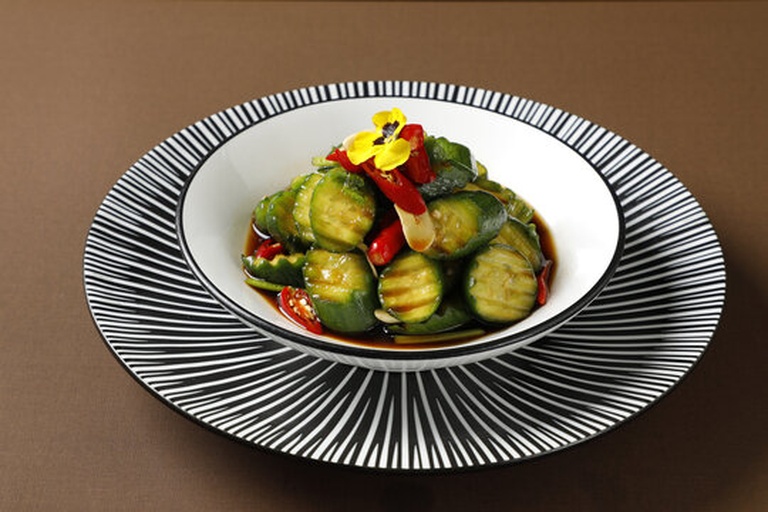 一盘晶莹剔透的腌制小黄瓜摆放在白色瓷盘中,黄瓜呈浅绿色,表面泛着光泽,点缀着红色小米辣和白色蒜片,旁边配有筷子和小碟,背景虚化 腌制完成的小黄瓜特写