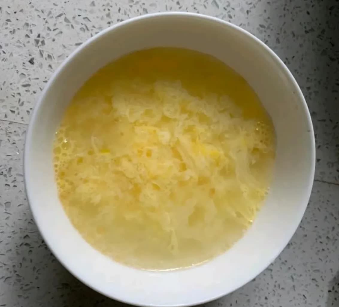 一碗刚冲好的鸡蛋羹,蛋液呈金黄色丝状凝结,漂浮在清汤中,表面滴有香油光泽,旁边放着瓷勺和调料瓶,背景为木质餐桌 开水冲蛋成品展示