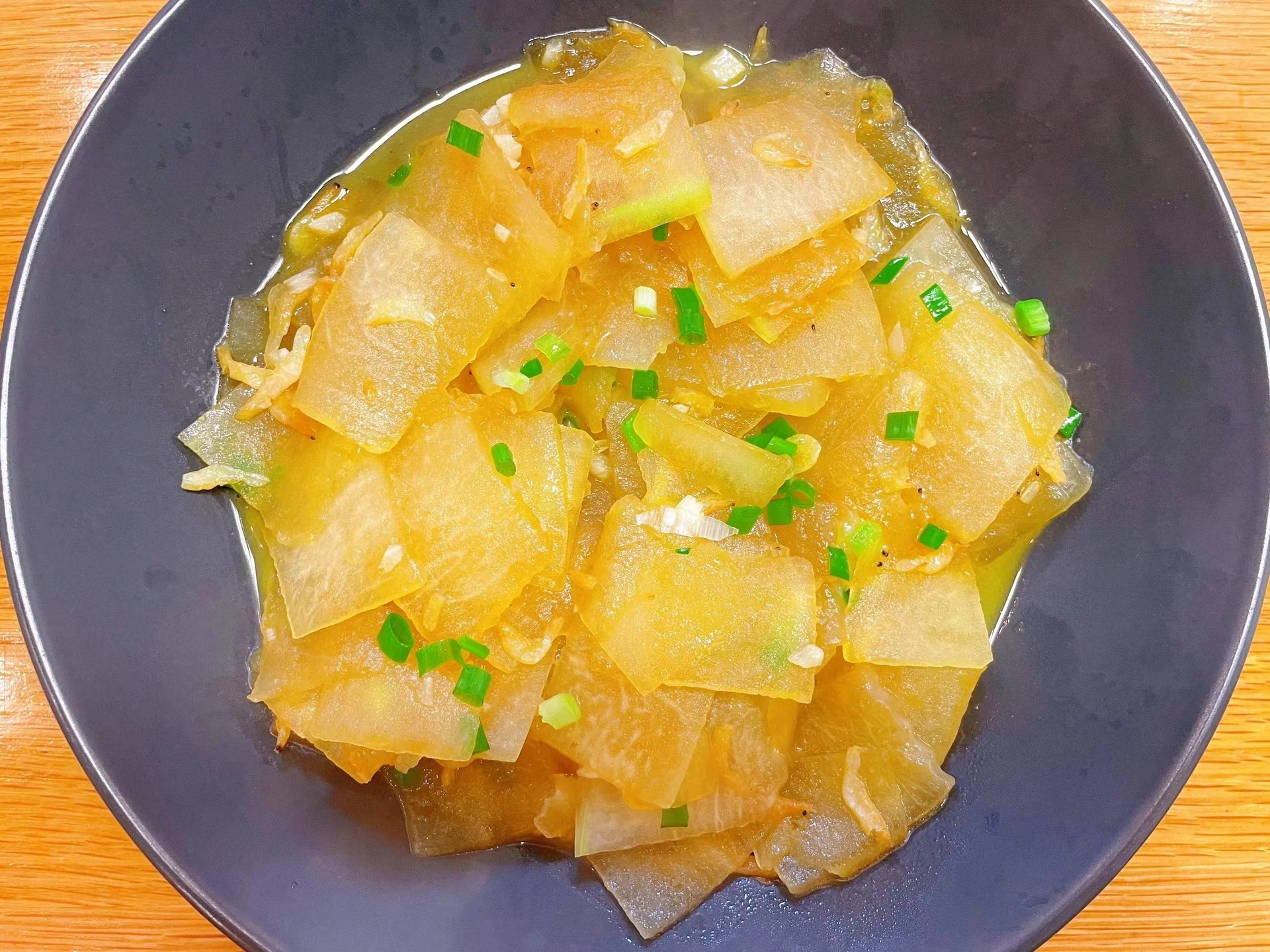 上海清炒冬瓜成品特写：晶莹透亮的冬瓜薄片整齐铺陈，表面覆薄层油润光泽，隐约可见浅琥珀色边缘，点缀翠绿葱丝与浅褐色香菇片