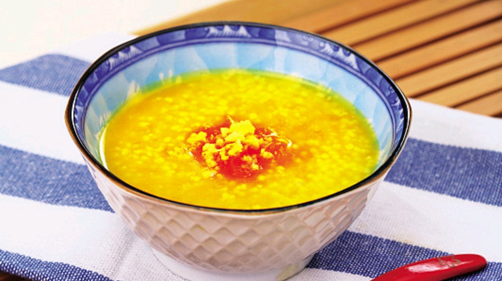 一碗热腾腾的红薯小米粥放置在木质餐桌上,橙黄色的红薯块均匀分布在金黄色的小米粥中,表面撒有少许枸杞,冒着淡淡热气,旁边配有竹筷和陶瓷勺 红薯小米粥盛在碗中的俯拍图