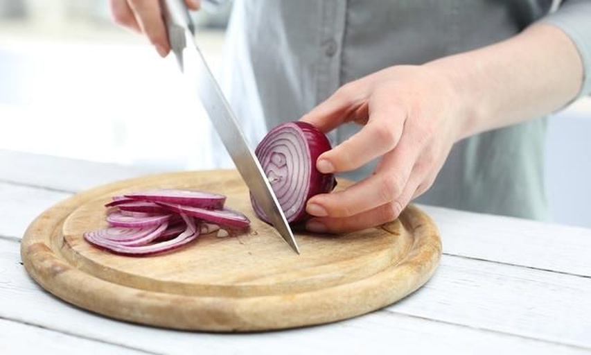 一位成年人正在厨房操作台上切洋葱,旁边放着蜂蜜罐、玻璃容器和量勺,阳光透过窗户洒入,展现温馨的家庭健康食品制备场景 家庭厨房中制作洋葱蜂蜜液的过程场景