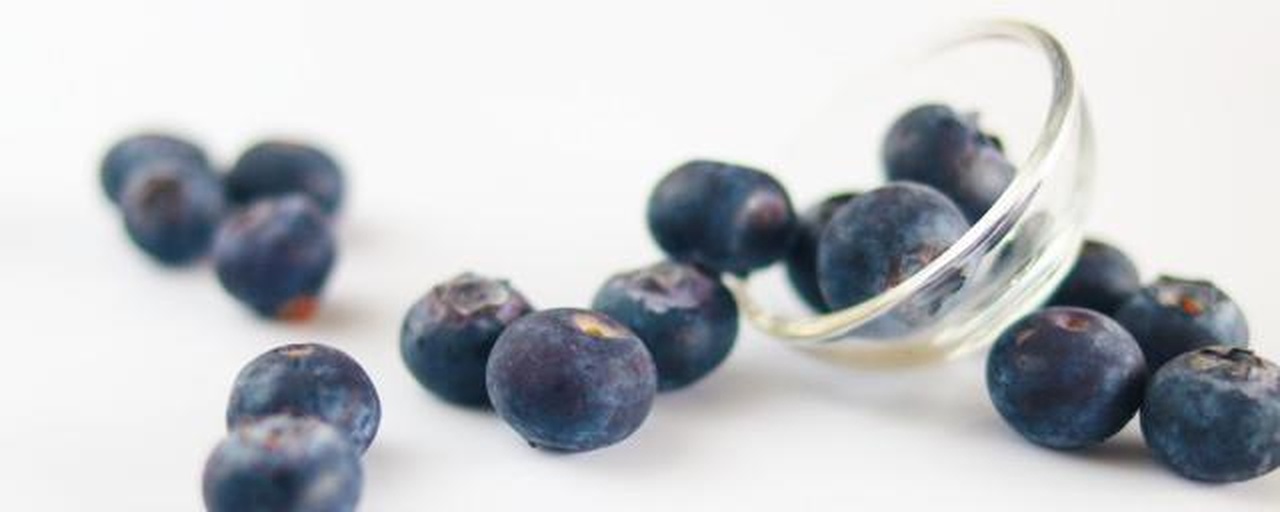 一个明亮现代的家庭厨房台面上,透明玻璃碗中盛有蓝色蓝莓,旁边是撒有白色粉末(小苏打)的容器,一只手正将蓝莓倒入滤网,水流清晰可见,背景有柠檬和绿色抹布,体现健康清洁氛围 家庭厨房中正在清洗蓝莓的场景