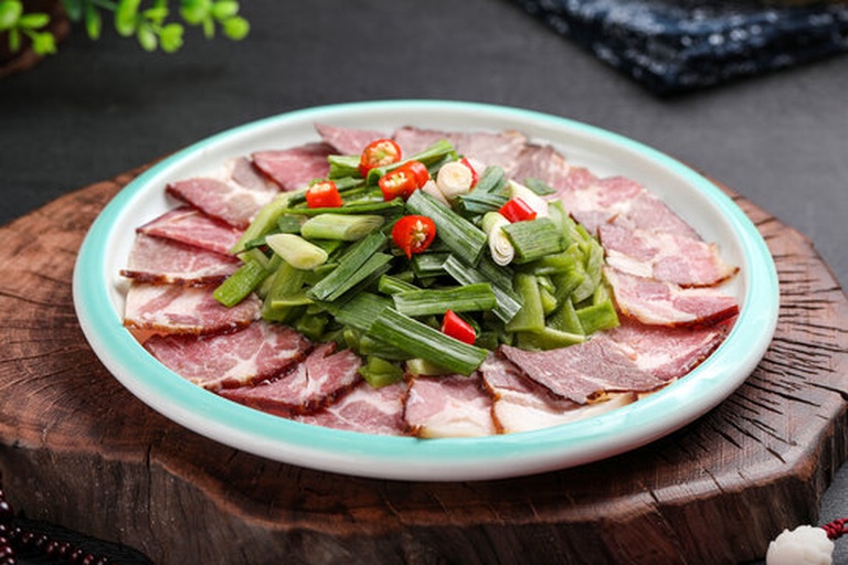 一位厨师正在厨房中翻炒锅中的食材,锅内清晰可见大量绿色扁平叶片,疑似蒜苗与红色腊肉混合翻炒,旁边砧板上放着未处理的蒜苗和葱,形成对比,展示实际烹饪中的使用区别 厨房应用场景图