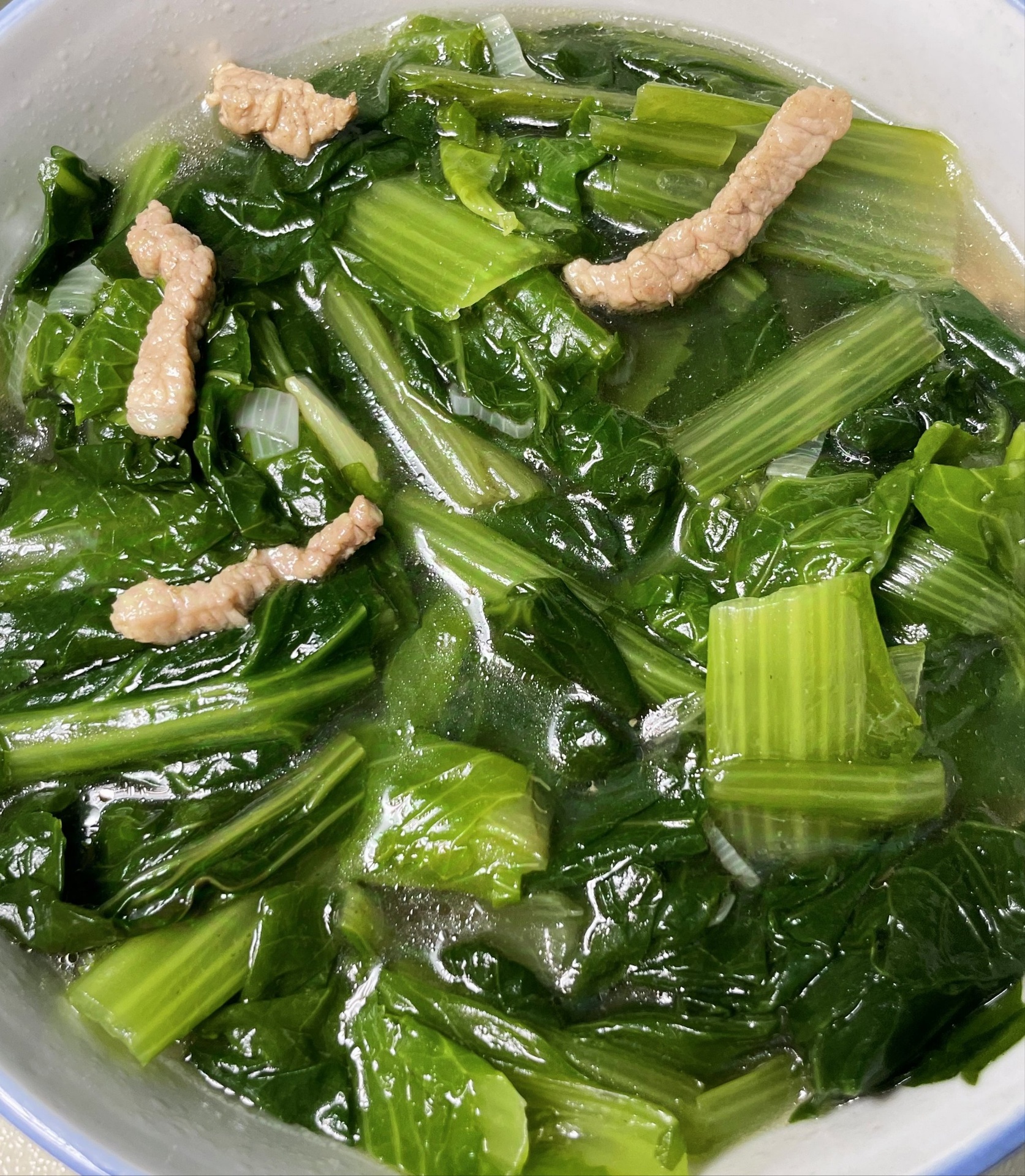 一碗热气腾腾的生菜瘦肉汤,汤色清澈微白,里面有薄切的猪肉片和舒展的绿色生菜叶,碗边配有小碟酱油和胡椒粉,背景为浅灰色餐布 生菜瘦肉汤特写